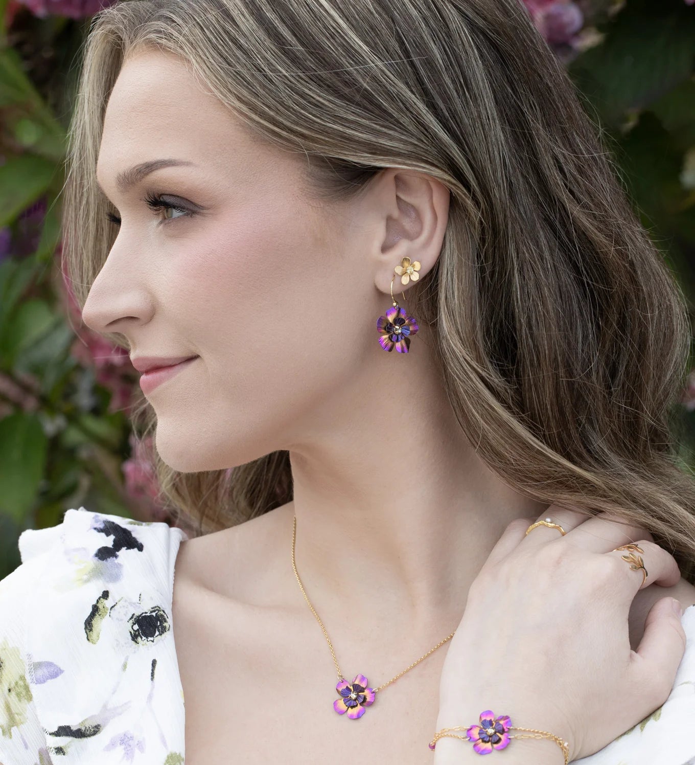 A woman with wavy blonde hair wears Holly Yashi’s Blissful Bloom Sparkling Fuchsia Earrings. She poses outdoors by pink hydrangeas, pairing the floral-shaped jewelry with a matching dress.