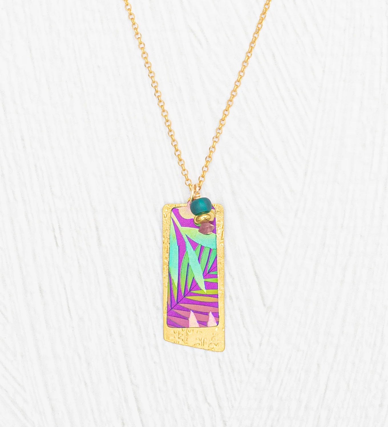 The Holly Yashi Davy Rectangle Two Layers Tropic Magenta Necklace features a gold chain with a rectangular pendant showcasing a hand-painted purple, teal, and green abstract leaf design, accented by a delicate turquoise bead and gold details on a white textured background—an elegant piece of jewelry.