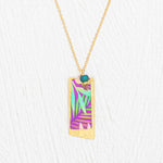 The Holly Yashi Davy Rectangle Two Layers Tropic Magenta Necklace features a gold chain with a rectangular pendant showcasing a hand-painted purple, teal, and green abstract leaf design, accented by a delicate turquoise bead and gold details on a white textured background—an elegant piece of jewelry.