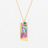 The Holly Yashi Davy Rectangle Two Layers Tropic Magenta Necklace features a gold chain with a rectangular pendant showcasing a hand-painted purple, teal, and green abstract leaf design, accented by a delicate turquoise bead and gold details on a white textured background—an elegant piece of jewelry.