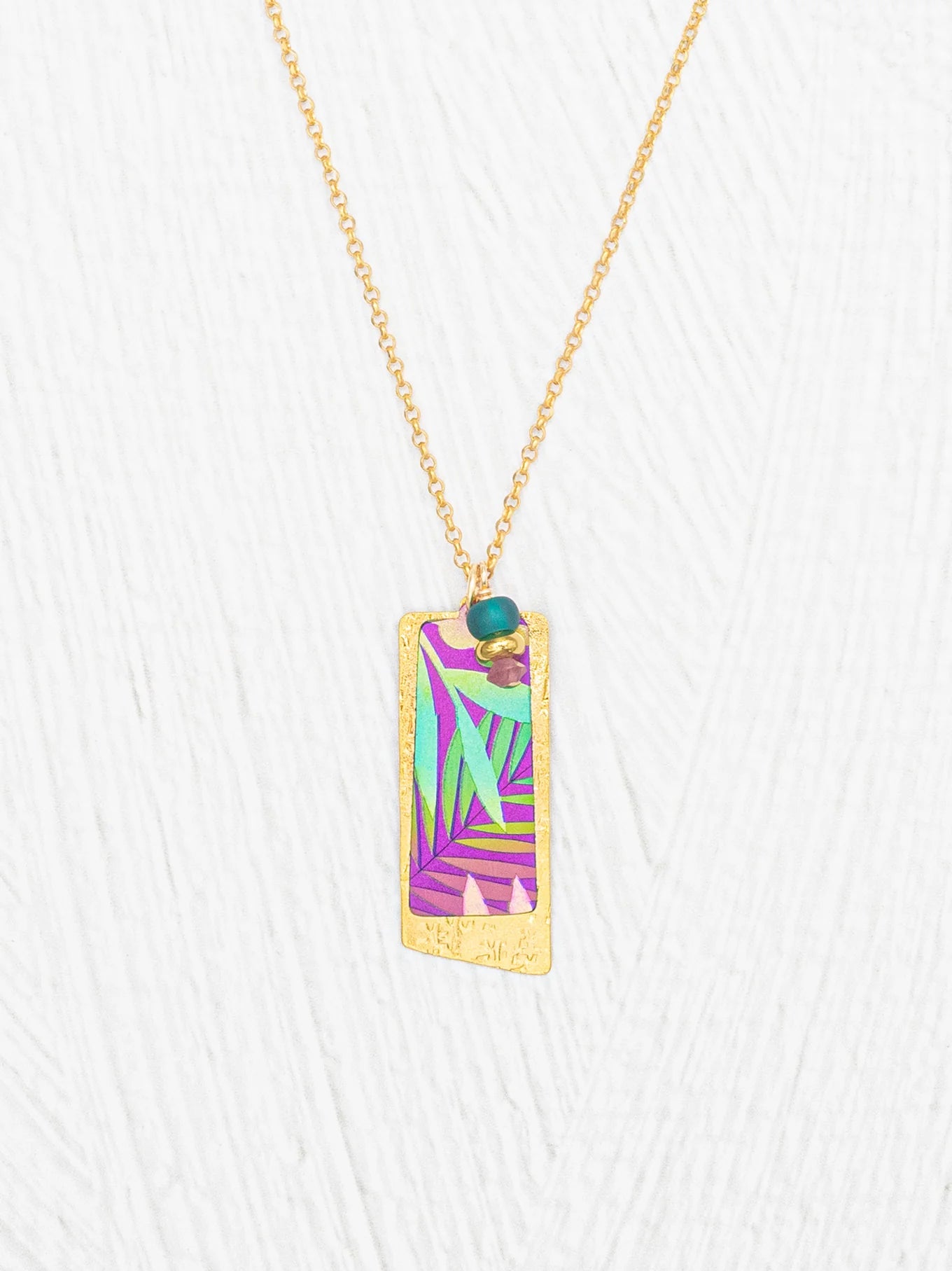 The Holly Yashi Davy Rectangle Two Layers Tropic Magenta Necklace features a gold chain with a rectangular pendant showcasing a hand-painted purple, teal, and green abstract leaf design, accented by a delicate turquoise bead and gold details on a white textured background—an elegant piece of jewelry.