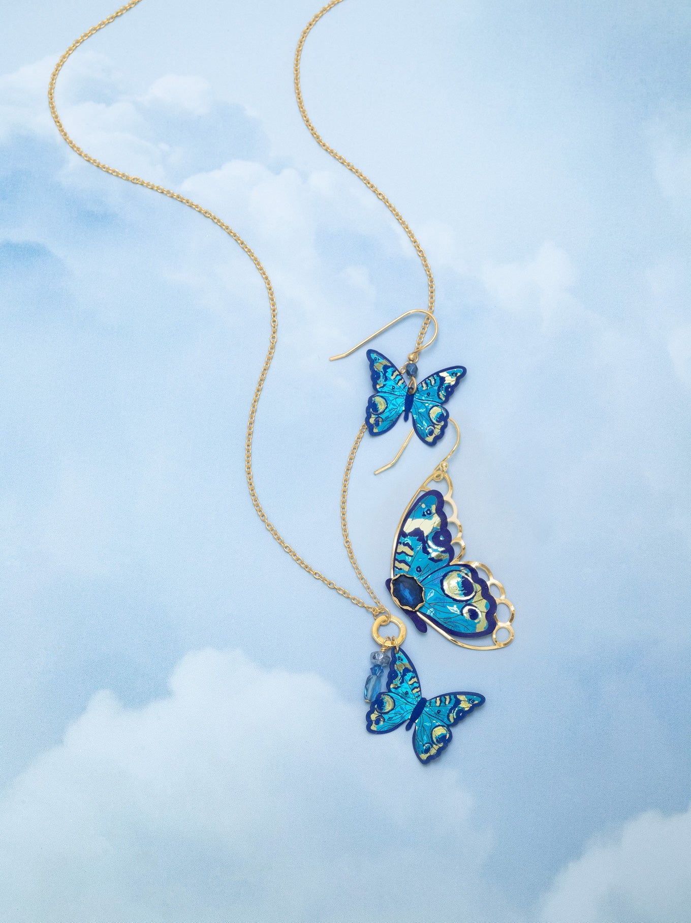 The Mariposa Butterfly Wing Blue Radiance Earrings by Holly Yashi are displayed on a light blue background with white clouds, resembling butterflies in the sky. Hypoallergenic and perfect for sensitive skin.