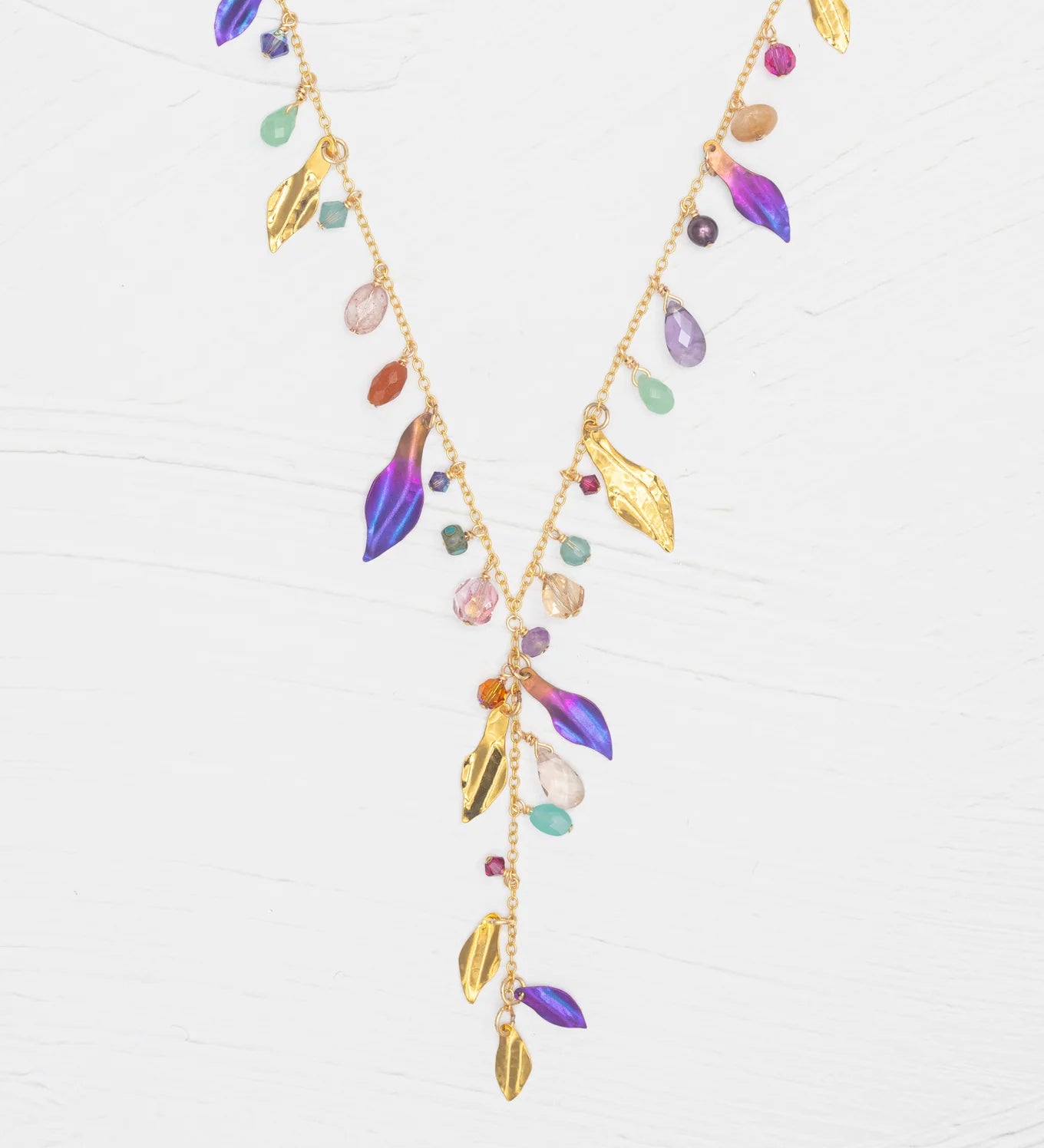 The Holly Yashi Calliope Tropic Punch Necklace showcases a gold-plated chain with gold leaves, colorful beads, and purple enamel niobium leaves in a V-shape on a white background.