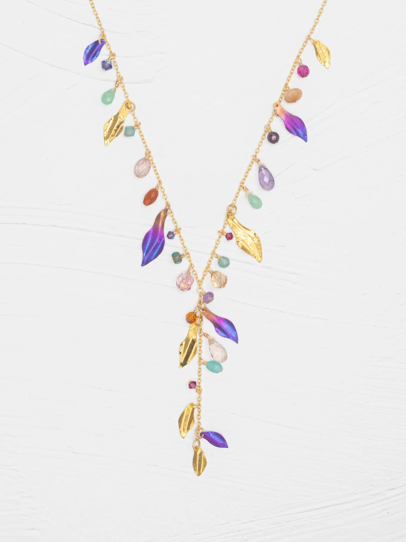 The Holly Yashi Calliope Tropic Punch Necklace showcases a gold-plated chain with gold leaves, colorful beads, and purple enamel niobium leaves in a V-shape on a white background.