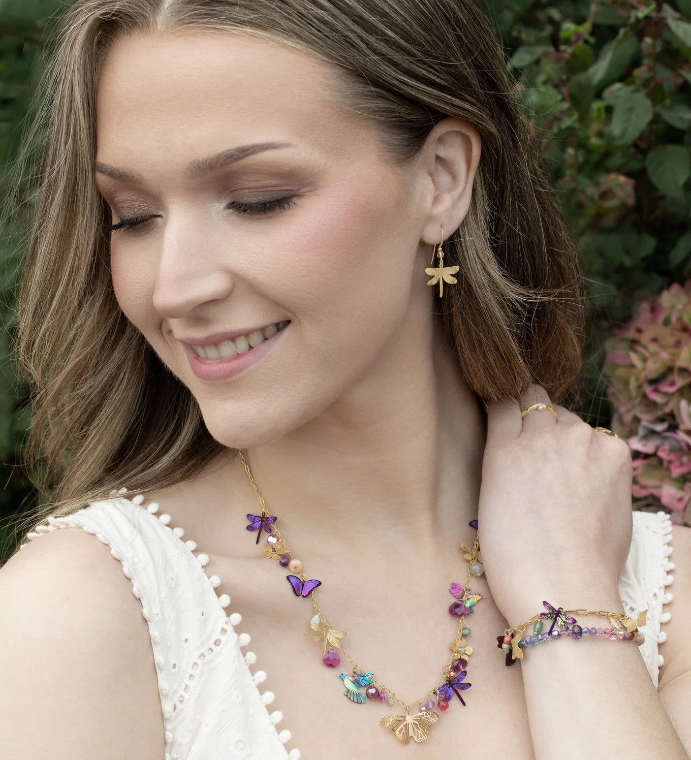 A woman wearing a white sleeveless top smiles, showcasing Holly Yashi’s Garden Party Gold Charms Necklace along with gold and colorful butterfly earrings and a vibrant niobium bracelet, with greenery and flowers softly blurred in the background.