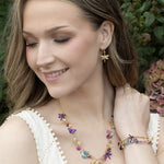 A woman wearing a white sleeveless top smiles, showcasing Holly Yashi’s Garden Party Gold Charms Necklace along with gold and colorful butterfly earrings and a vibrant niobium bracelet, with greenery and flowers softly blurred in the background.