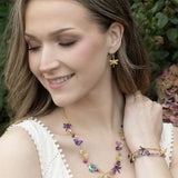 A woman wearing a white sleeveless top smiles, showcasing Holly Yashi’s Garden Party Gold Charms Necklace along with gold and colorful butterfly earrings and a vibrant niobium bracelet, with greenery and flowers softly blurred in the background.