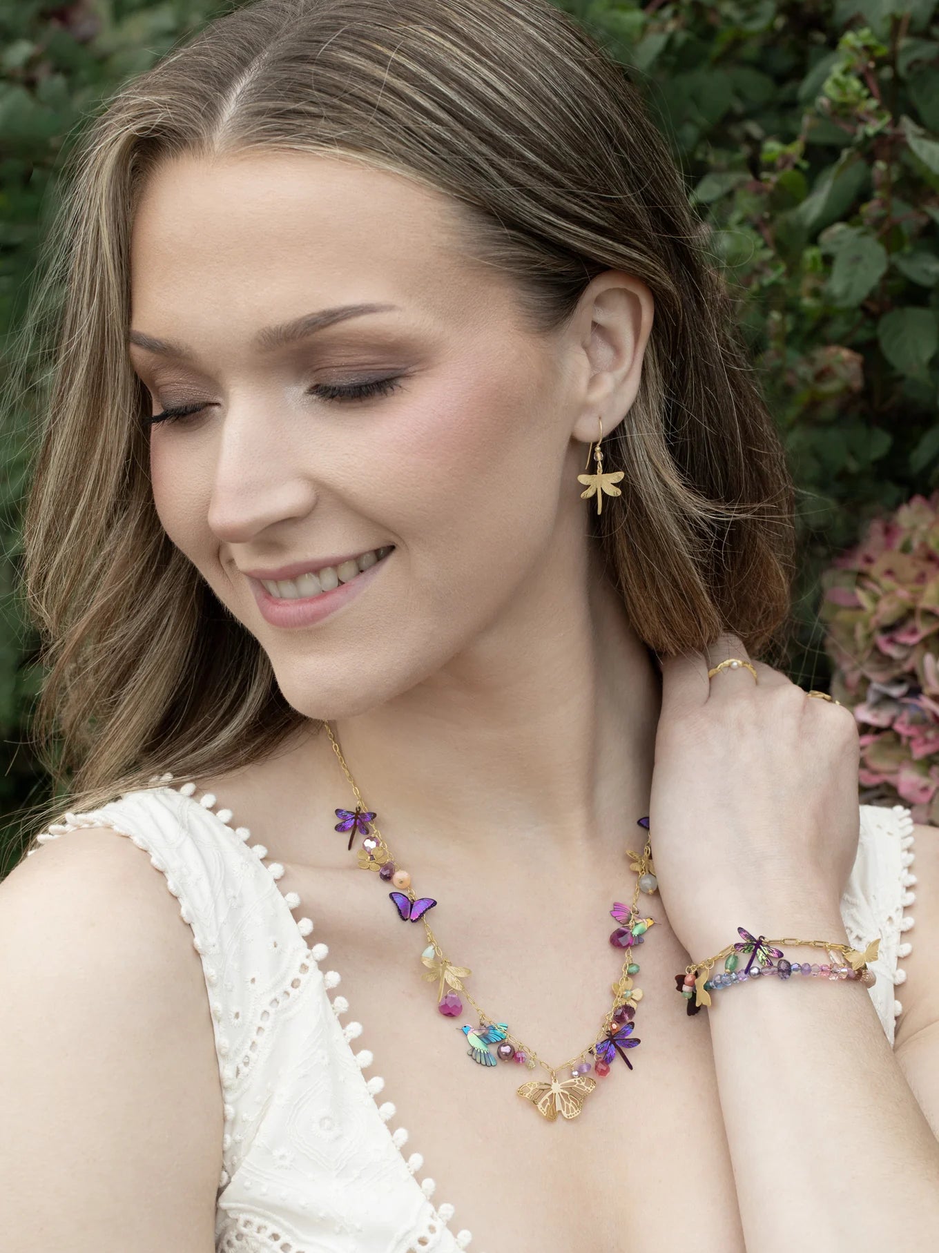 A woman wearing a white sleeveless top smiles, showcasing Holly Yashi’s Garden Party Gold Charms Necklace along with gold and colorful butterfly earrings and a vibrant niobium bracelet, with greenery and flowers softly blurred in the background.