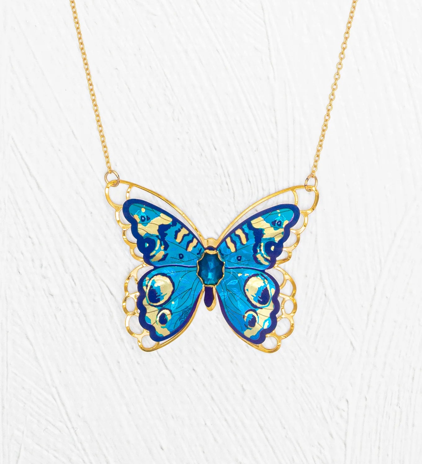 The Holly Yashi NK: Mariposa Butterfly Blue Radiance features a gold butterfly necklace with a large blue enamel pendant, intricate detailing, and an adjustable chain, shown against a white textured background.