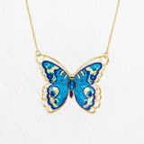 The Holly Yashi NK: Mariposa Butterfly Blue Radiance features a gold butterfly necklace with a large blue enamel pendant, intricate detailing, and an adjustable chain, shown against a white textured background.