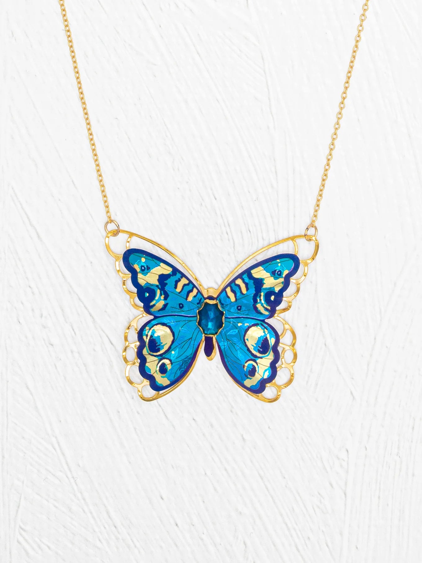 The Holly Yashi NK: Mariposa Butterfly Blue Radiance features a gold butterfly necklace with a large blue enamel pendant, intricate detailing, and an adjustable chain, shown against a white textured background.
