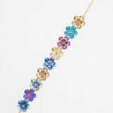 The Blissful Bouquet Multicolor Bracelet by Holly Yashi is a gold chain adorned with iridescent niobium flowers, displayed on a white textured background. Handcrafted, this vibrant piece brings colorful charm to any outfit.
