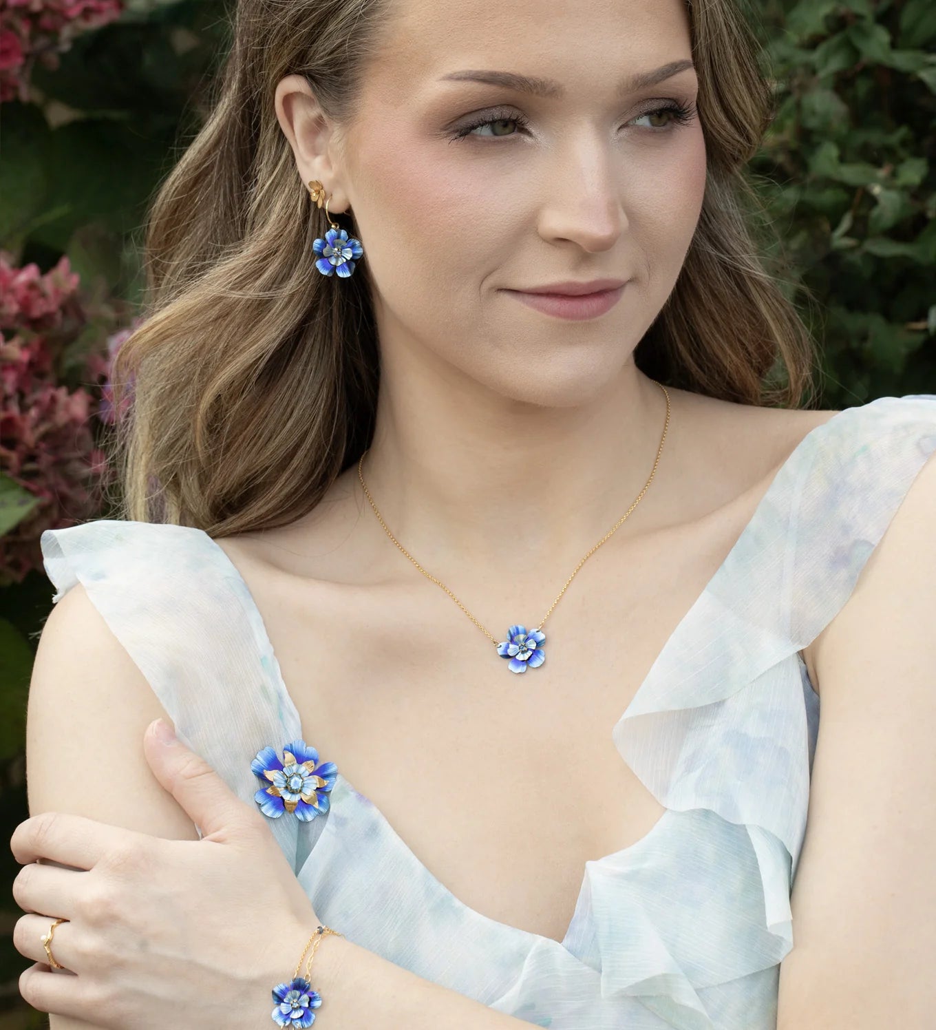 A woman with wavy light brown hair wears a light blue ruffled dress and matching blue flower jewelry, including Holly Yashi’s Blissful Bloom Bonnie Blue Necklace, while standing outdoors near pink flowers and greenery.