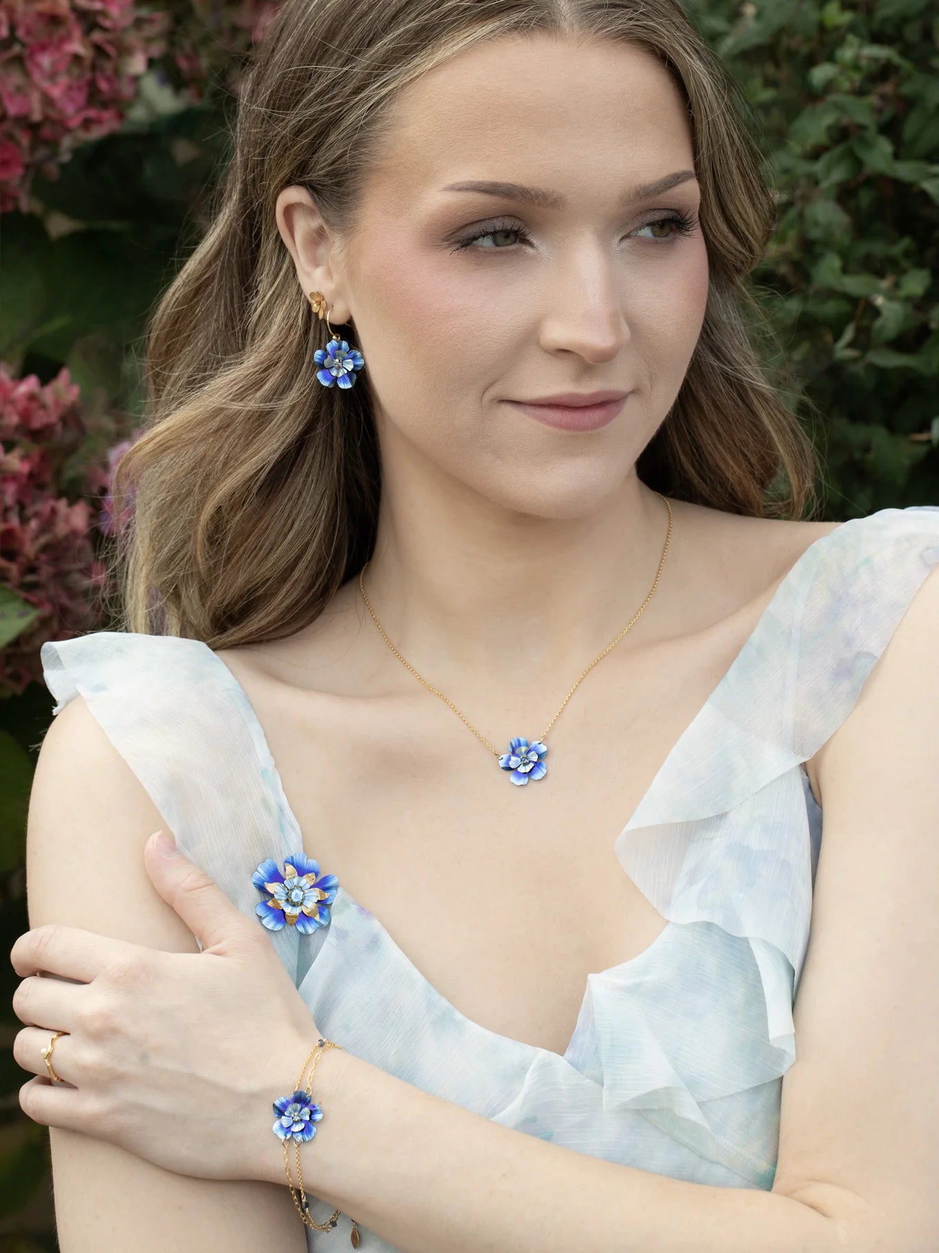 A woman with wavy light brown hair wears a light blue ruffled dress and matching blue flower jewelry, including Holly Yashi’s Blissful Bloom Bonnie Blue Necklace, while standing outdoors near pink flowers and greenery.