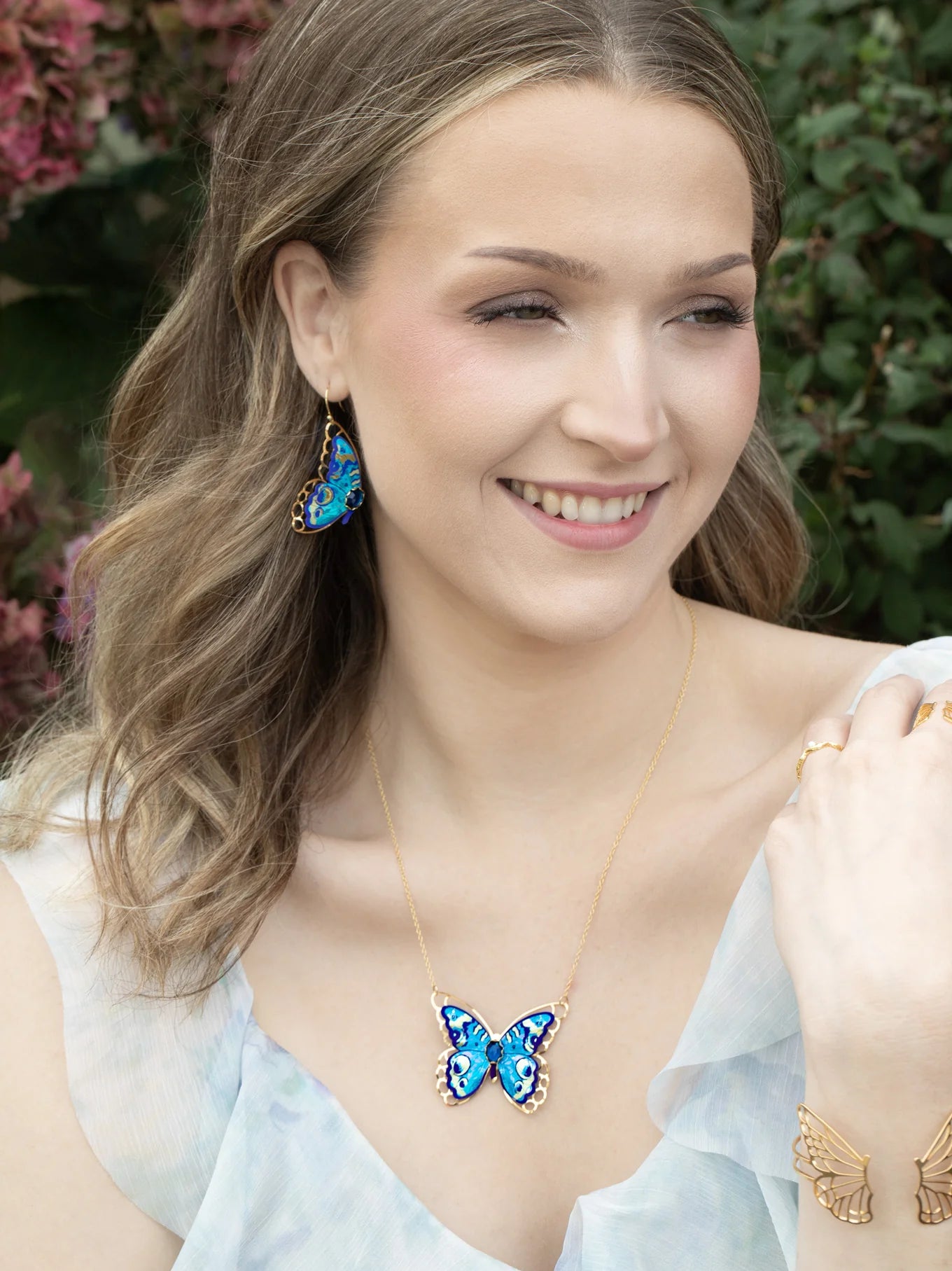 A woman with wavy light brown hair smiles outdoors, wearing Holly Yashi's Mariposa Butterfly Wing Blue Radiance Earrings and a matching butterfly necklace. She also wears a delicate bracelet and pale, flowy dress amid greenery and pink flowers.