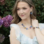 A woman in a light, ruffled dress poses by blooming hydrangeas, wearing handcrafted Holly Yashi jewelry—a matching set of blue and purple floral pieces, including the Blissful Bouquet Multicolor Bracelet. She smiles softly and looks to the side.