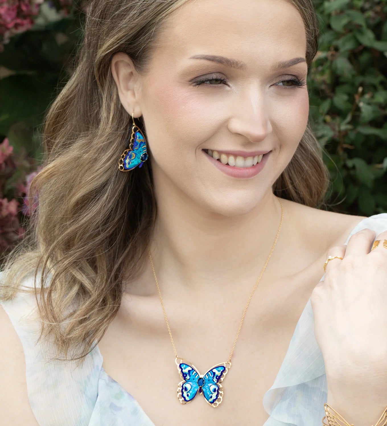 A woman with wavy light brown hair smiles outdoors, wearing the Holly Yashi NK: Mariposa Butterfly Blue Radiance necklace, matching earrings, and a butterfly bracelet. Pink flowers and greenery form a charming backdrop.