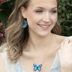 A woman with wavy light brown hair smiles outdoors, wearing the Holly Yashi NK: Mariposa Butterfly Blue Radiance necklace, matching earrings, and a butterfly bracelet. Pink flowers and greenery form a charming backdrop.