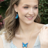 A woman with wavy light brown hair smiles outdoors, wearing the Holly Yashi NK: Mariposa Butterfly Blue Radiance necklace, matching earrings, and a butterfly bracelet. Pink flowers and greenery form a charming backdrop.