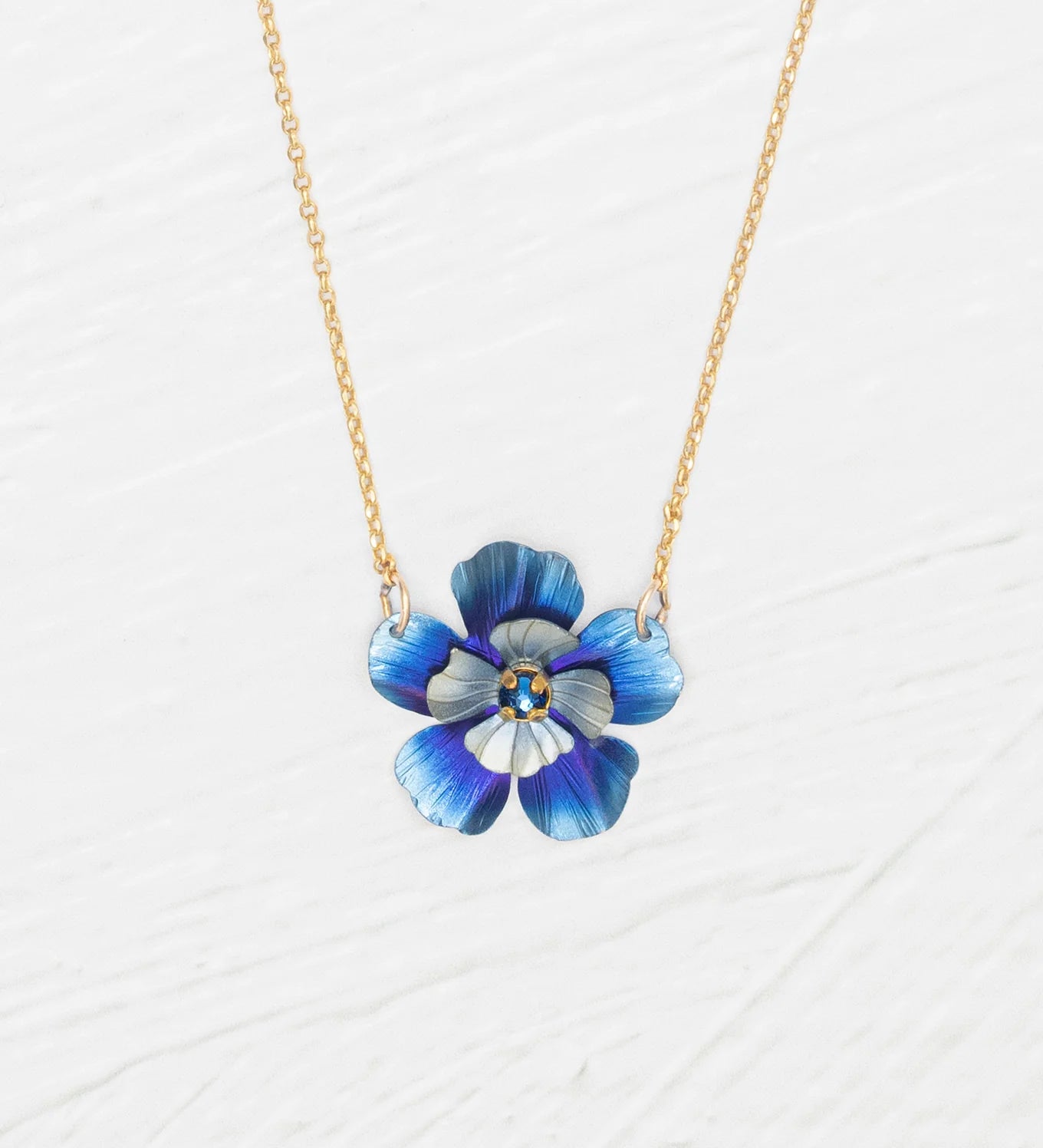 The Holly Yashi Blissful Bloom Bonnie Blue Necklace features a gold adjustable chain with a blue flower pendant, detailed petals, and a yellow center, set against a white textured background.