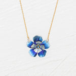 The Holly Yashi Blissful Bloom Bonnie Blue Necklace features a gold adjustable chain with a blue flower pendant, detailed petals, and a yellow center, set against a white textured background.