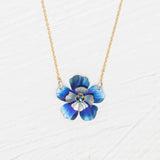 The Holly Yashi Blissful Bloom Bonnie Blue Necklace features a gold adjustable chain with a blue flower pendant, detailed petals, and a yellow center, set against a white textured background.