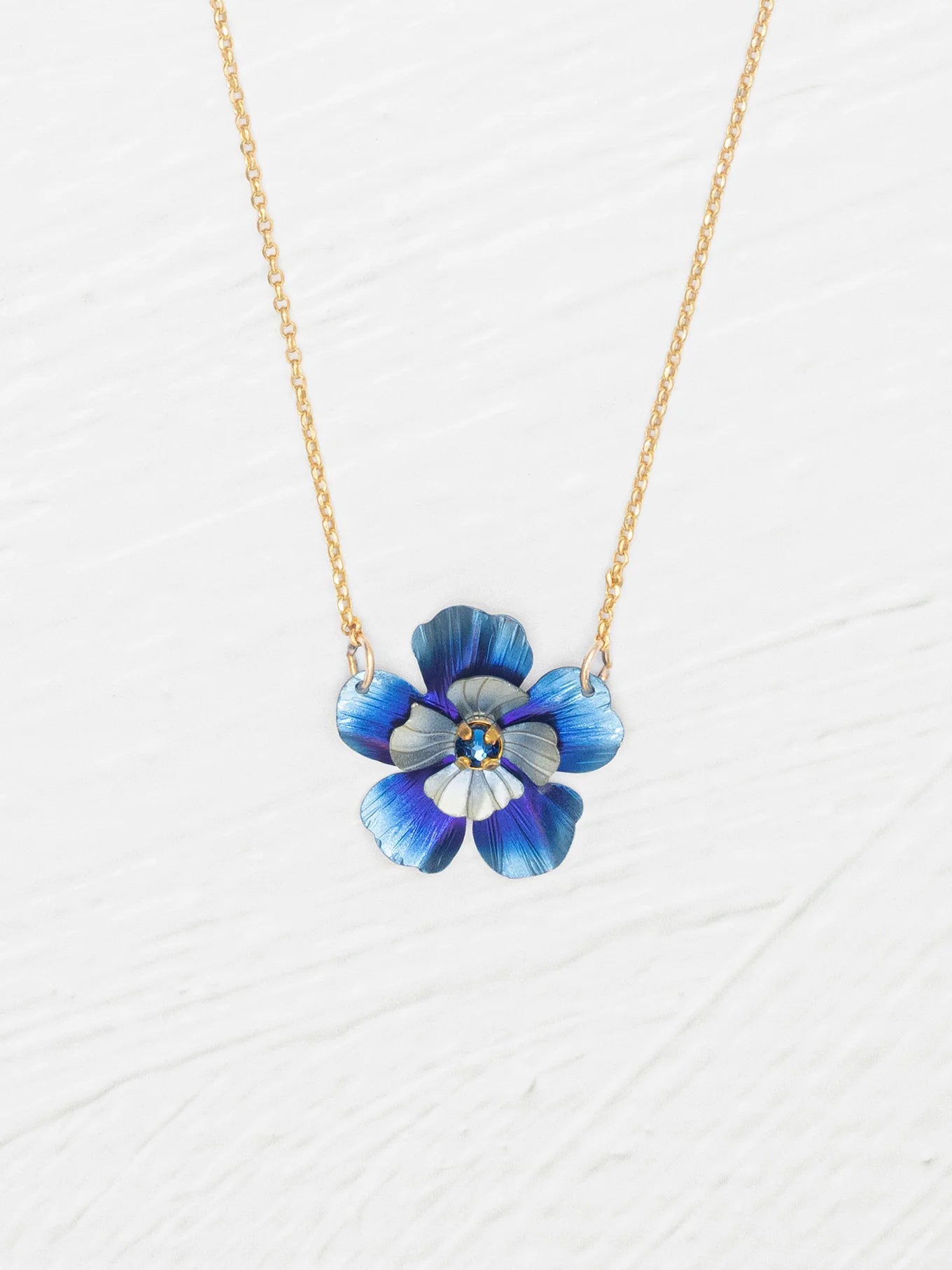 The Holly Yashi Blissful Bloom Bonnie Blue Necklace features a gold adjustable chain with a blue flower pendant, detailed petals, and a yellow center, set against a white textured background.