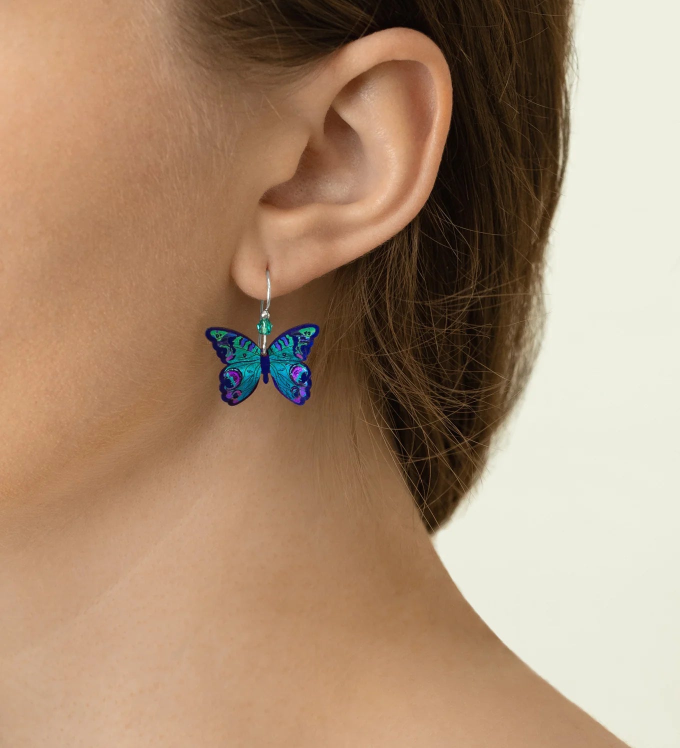 Close-up of a person with light skin and brown hair wearing Holly Yashi's ER: Mariposa Butterfly Blue Radiance earrings—hypoallergenic, gold-filled, and adorned in vibrant blue, green, and purple tones. Perfect for sensitive ears.