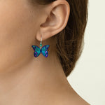 Close-up of a person with light skin and brown hair wearing Holly Yashi's ER: Mariposa Butterfly Blue Radiance earrings—hypoallergenic, gold-filled, and adorned in vibrant blue, green, and purple tones. Perfect for sensitive ears.