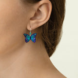 Close-up of a person with light skin and brown hair wearing Holly Yashi's ER: Mariposa Butterfly Blue Radiance earrings—hypoallergenic, gold-filled, and adorned in vibrant blue, green, and purple tones. Perfect for sensitive ears.