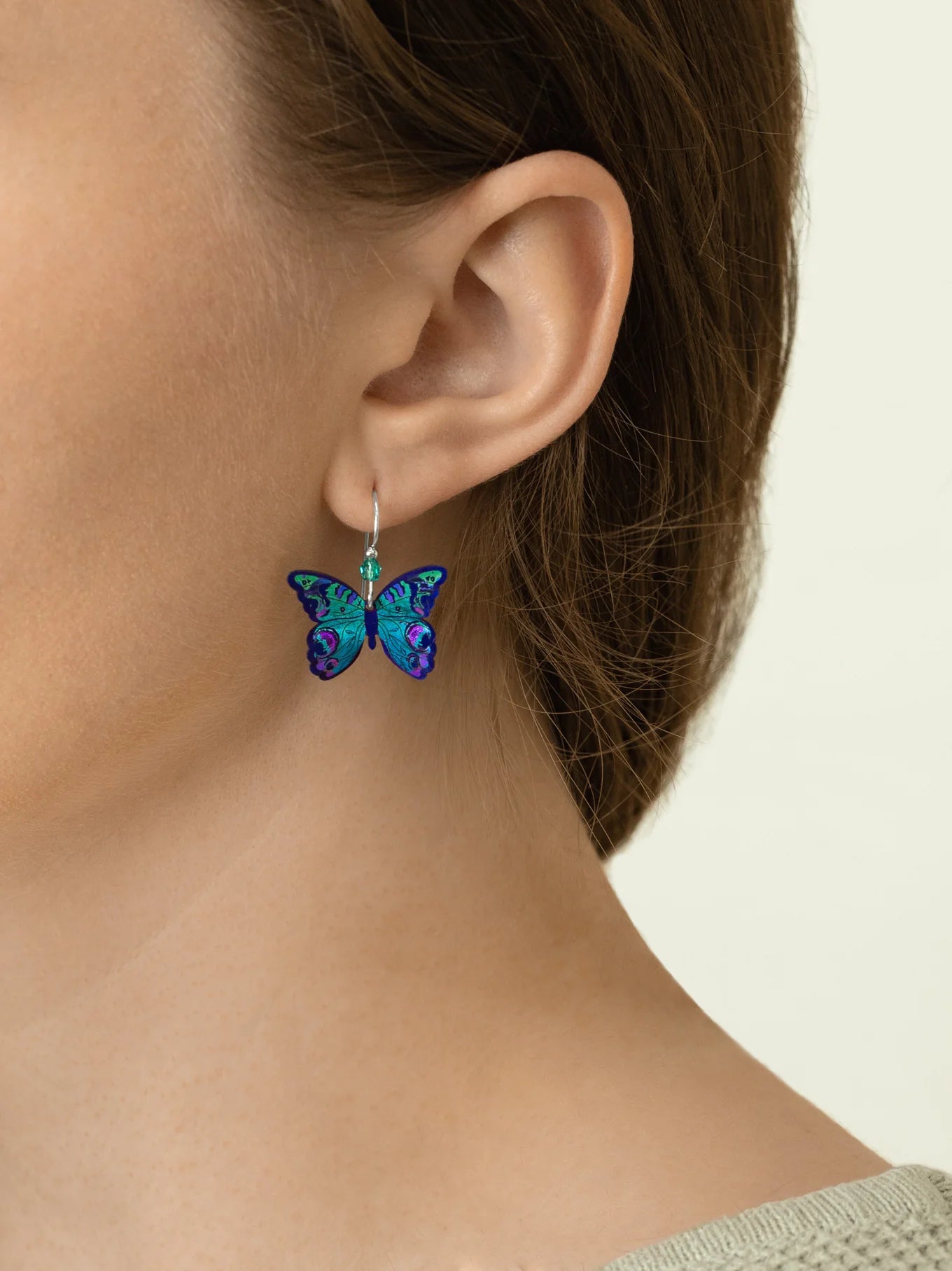 Close-up of a person with light skin and brown hair wearing Holly Yashi's ER: Mariposa Butterfly Blue Radiance earrings—hypoallergenic, gold-filled, and adorned in vibrant blue, green, and purple tones. Perfect for sensitive ears.