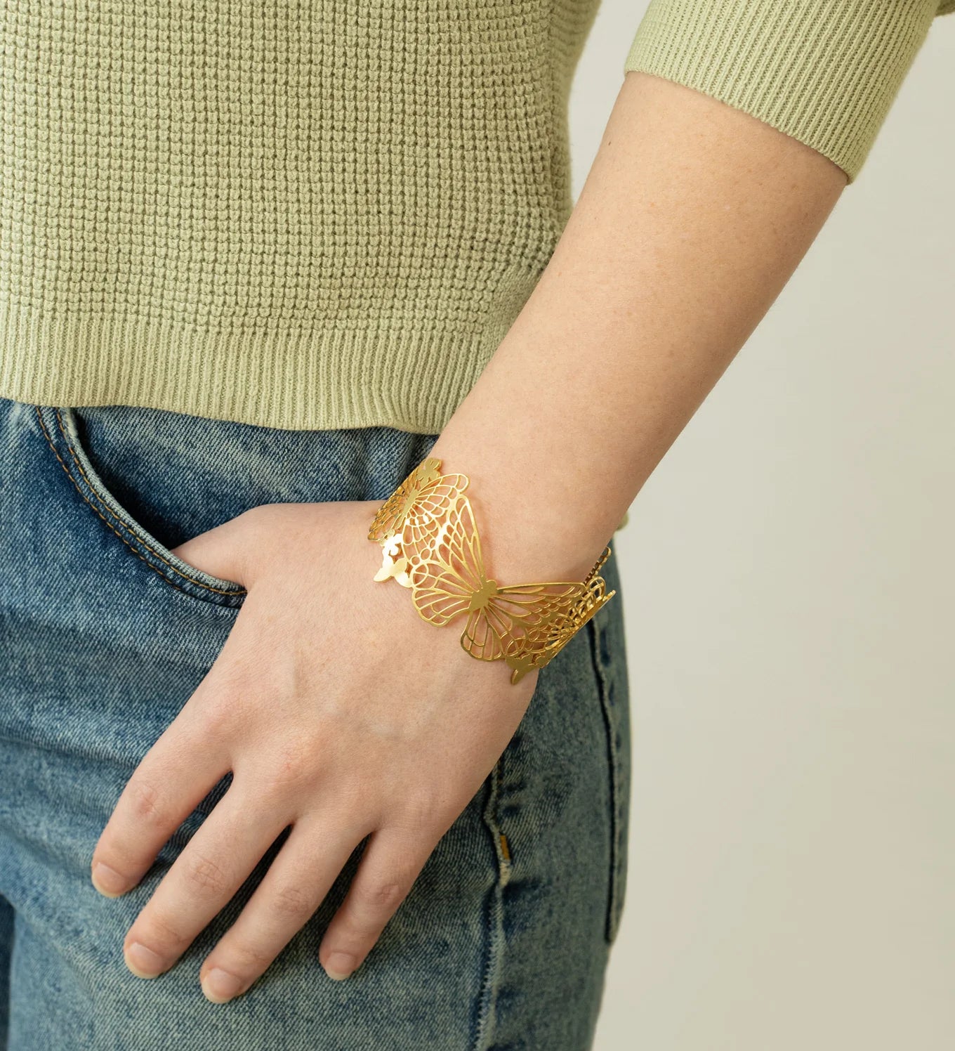 A person wearing blue jeans and a light green sweater has an eye-catching Holly Yashi Butterfly Garden Gold Cuff on their wrist, with their hand relaxed in their pocket.