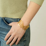 A person wearing blue jeans and a light green sweater has an eye-catching Holly Yashi Butterfly Garden Gold Cuff on their wrist, with their hand relaxed in their pocket.