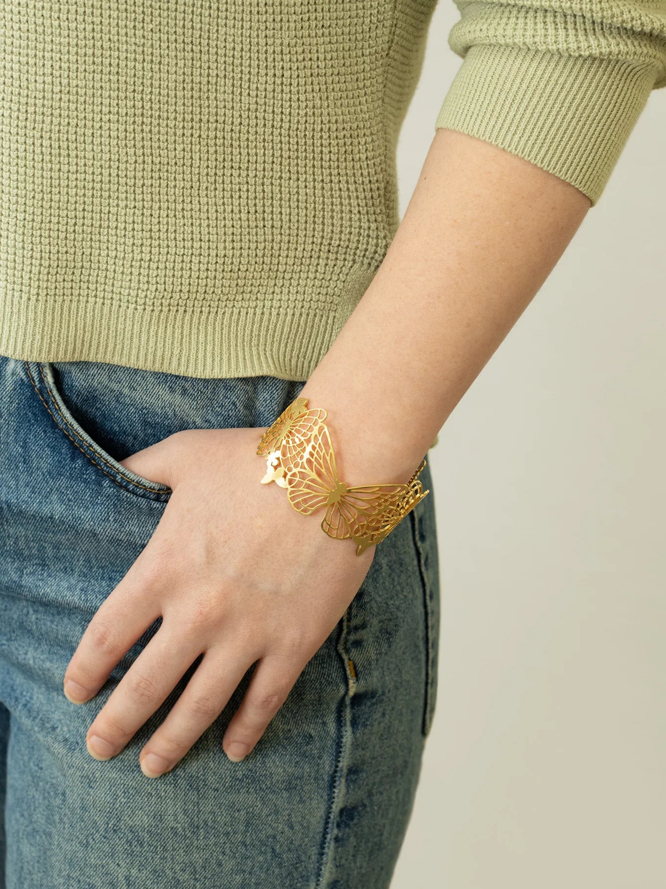 A person wearing blue jeans and a light green sweater has an eye-catching Holly Yashi Butterfly Garden Gold Cuff on their wrist, with their hand relaxed in their pocket.