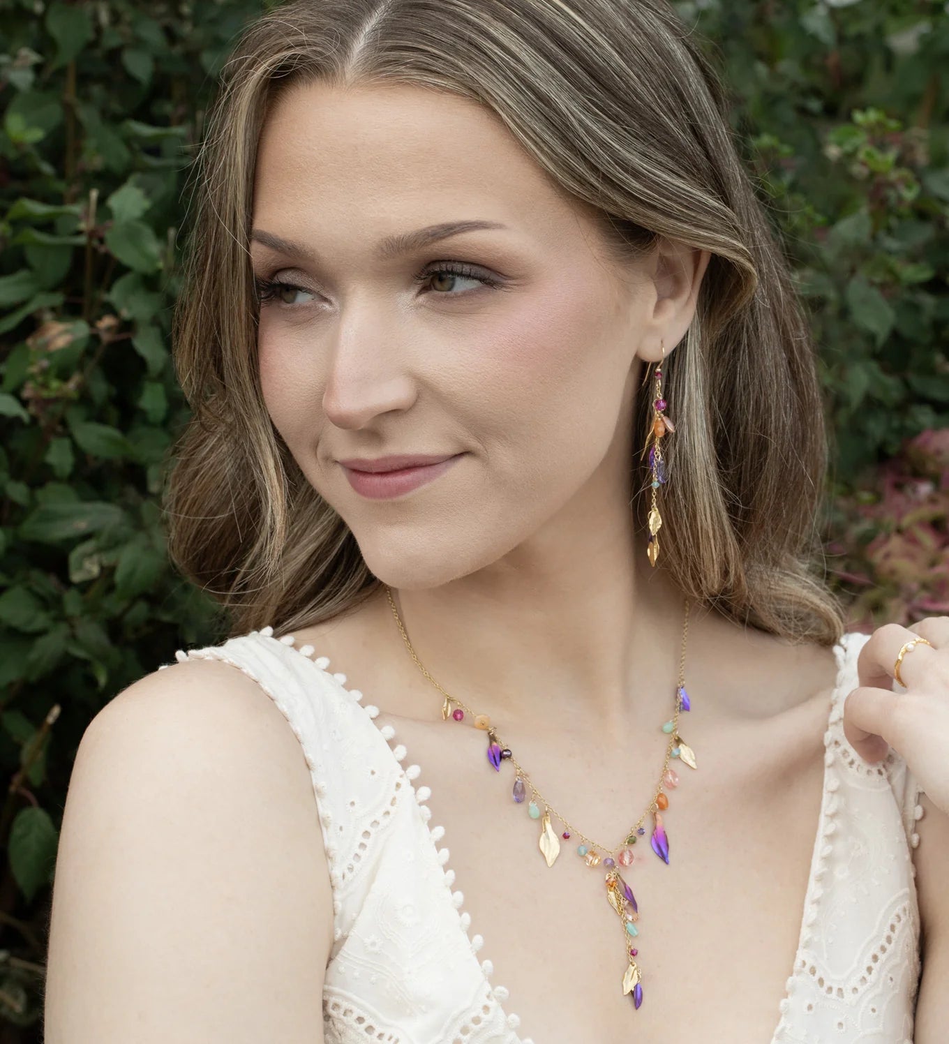A woman with wavy light brown hair wears a white sleeveless top and colorful gold-plated jewelry, including drop earrings and the Holly Yashi Calliope Tropic Punch Necklace with niobium leaves, while posing outdoors amid green foliage.