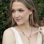A woman with wavy light brown hair wears a white sleeveless top and colorful gold-plated jewelry, including drop earrings and the Holly Yashi Calliope Tropic Punch Necklace with niobium leaves, while posing outdoors amid green foliage.