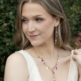 A woman with wavy light brown hair wears a white sleeveless top and colorful gold-plated jewelry, including drop earrings and the Holly Yashi Calliope Tropic Punch Necklace with niobium leaves, while posing outdoors amid green foliage.