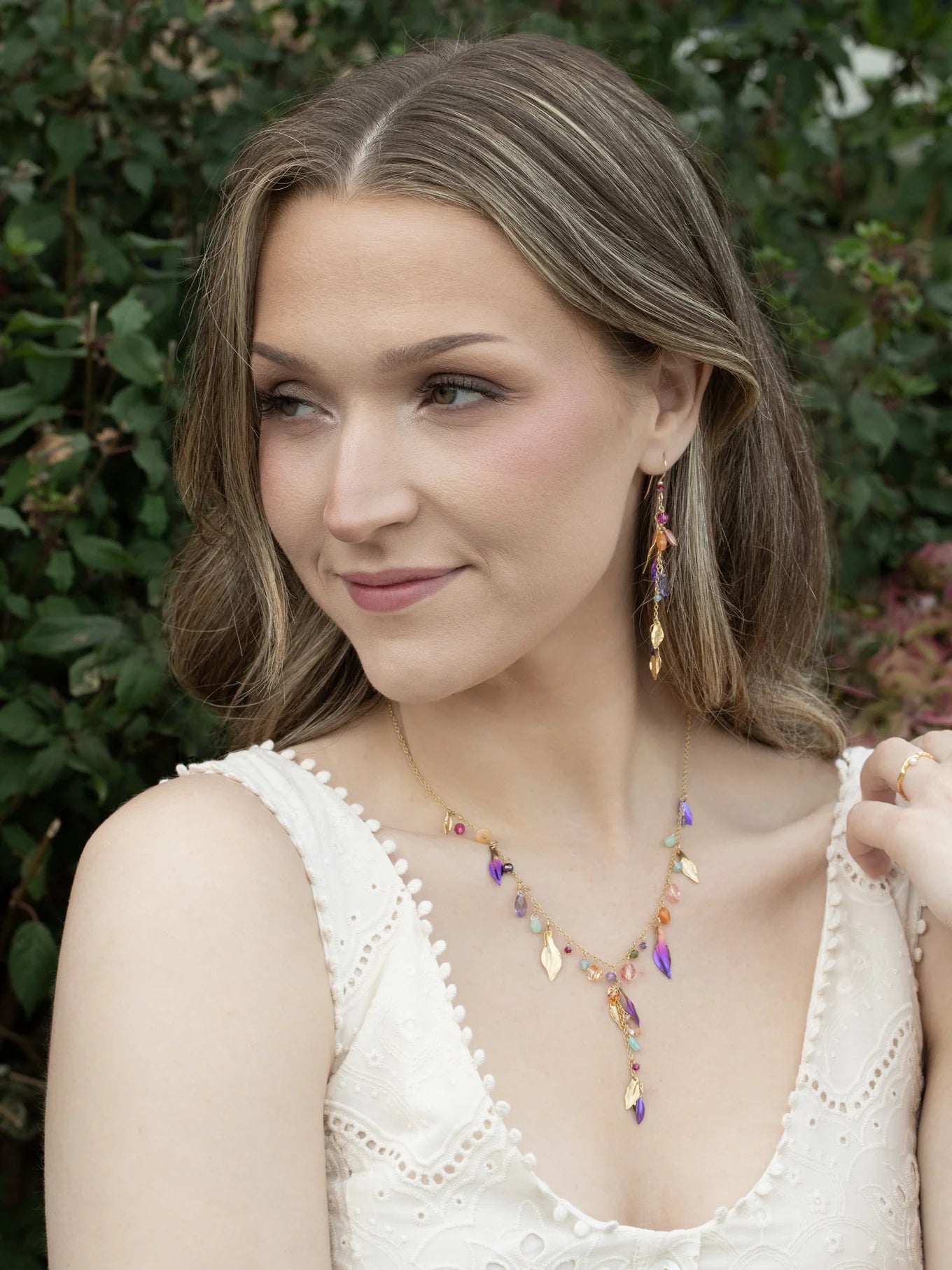 A woman with wavy light brown hair wears a white sleeveless top and colorful gold-plated jewelry, including drop earrings and the Holly Yashi Calliope Tropic Punch Necklace with niobium leaves, while posing outdoors amid green foliage.