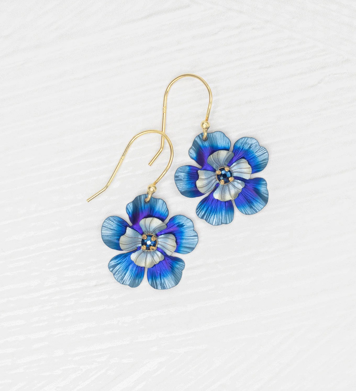 Discover Holly Yashi’s Blissful Bloom Bonnie Blue Earrings: gold hook, hypo-allergenic, featuring blue, purple, and white enamel flowers with yellow and white centers, beautifully displayed on a textured white surface.