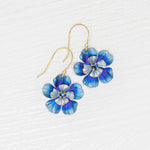 Discover Holly Yashi’s Blissful Bloom Bonnie Blue Earrings: gold hook, hypo-allergenic, featuring blue, purple, and white enamel flowers with yellow and white centers, beautifully displayed on a textured white surface.
