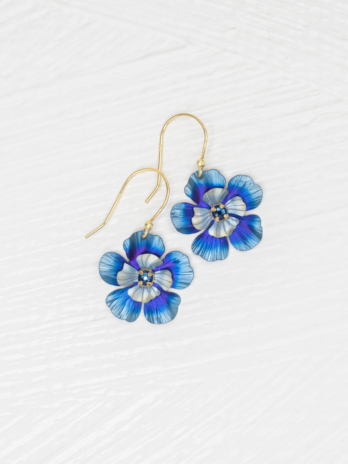 Discover Holly Yashi’s Blissful Bloom Bonnie Blue Earrings: gold hook, hypo-allergenic, featuring blue, purple, and white enamel flowers with yellow and white centers, beautifully displayed on a textured white surface.