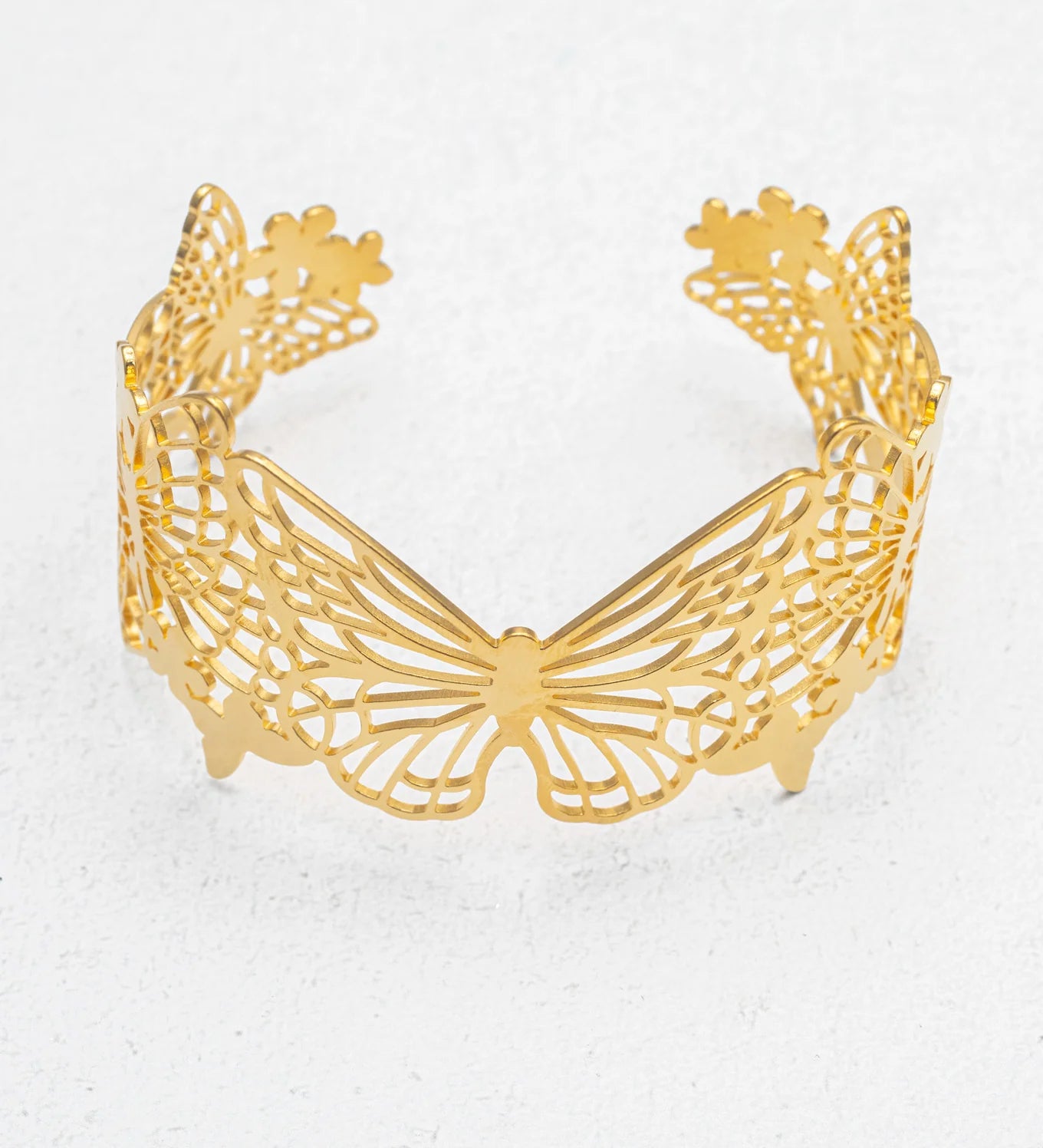 The Butterfly Garden Gold Cuff by Holly Yashi features an intricate cutout butterfly design, creating an elegant wing motif that stands out as a striking statement piece, showcased upright on a lightly textured surface.