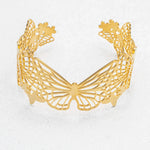 The Butterfly Garden Gold Cuff by Holly Yashi features an intricate cutout butterfly design, creating an elegant wing motif that stands out as a striking statement piece, showcased upright on a lightly textured surface.