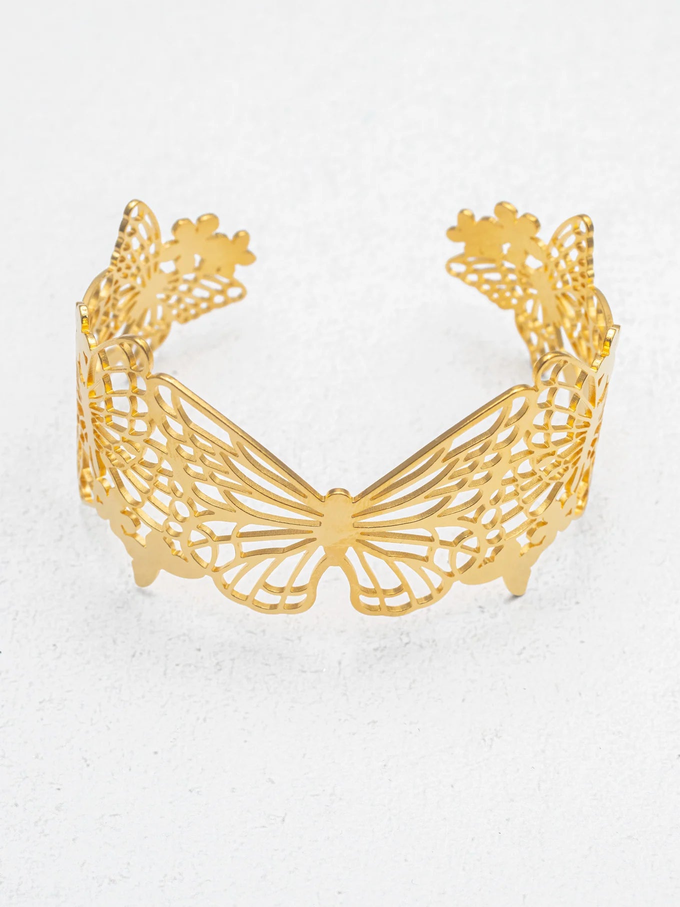 The Butterfly Garden Gold Cuff by Holly Yashi features an intricate cutout butterfly design, creating an elegant wing motif that stands out as a striking statement piece, showcased upright on a lightly textured surface.