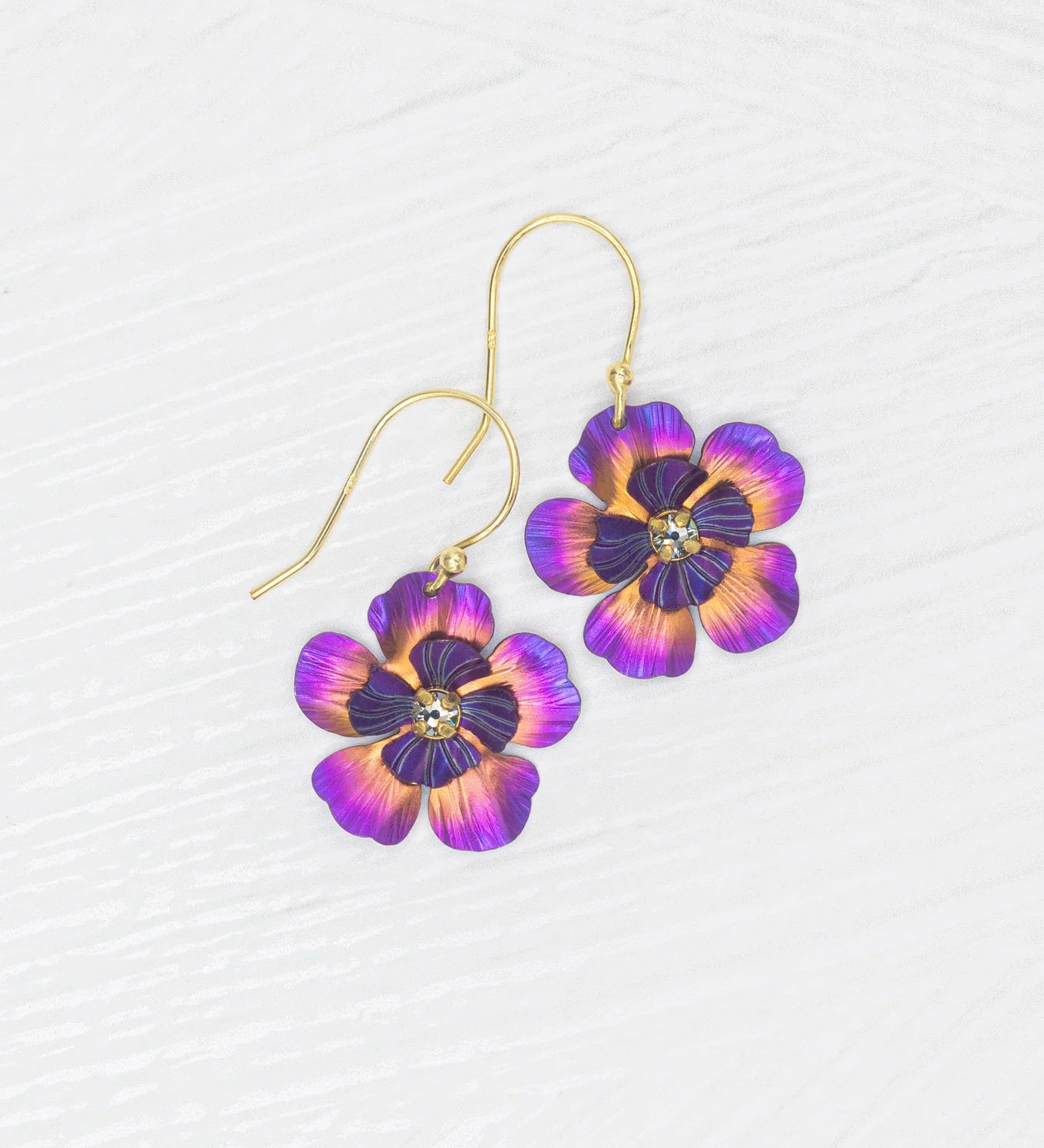The Holly Yashi Blissful Bloom Sparkling Fuchsia Earrings feature gold-filled French wires, purple and orange flower-shaped pendants, and a rhinestone center, displayed on a white textured background.