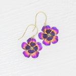The Holly Yashi Blissful Bloom Sparkling Fuchsia Earrings feature gold-filled French wires, purple and orange flower-shaped pendants, and a rhinestone center, displayed on a white textured background.