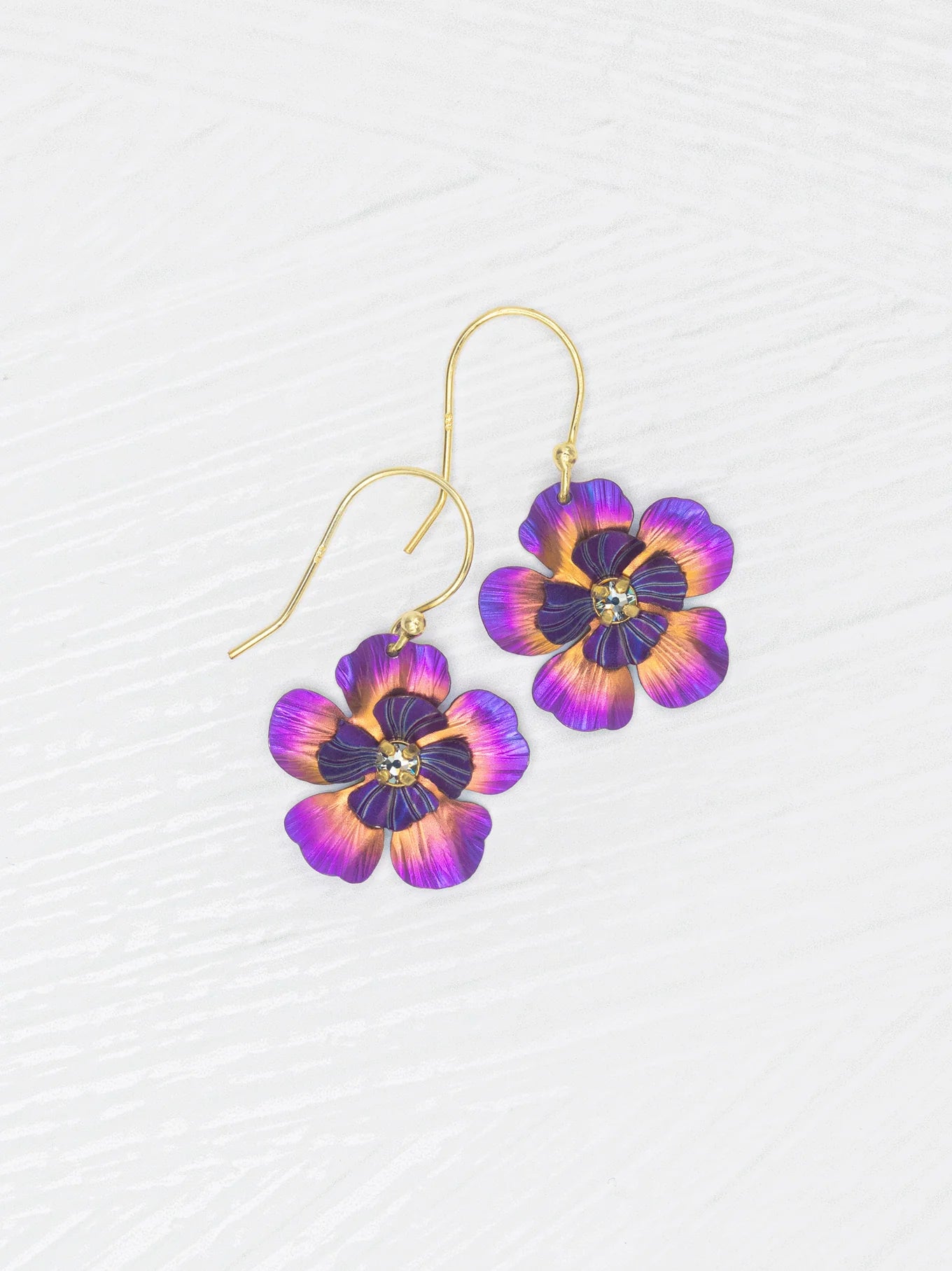 The Holly Yashi Blissful Bloom Sparkling Fuchsia Earrings feature gold-filled French wires, purple and orange flower-shaped pendants, and a rhinestone center, displayed on a white textured background.