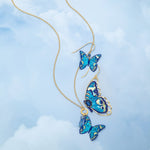 The Holly Yashi NK: Mariposa Butterfly Blue Radiance necklace features three gold butterflies with blue pendants on an adjustable chain, arranged against a soft sky background for an elegant look.