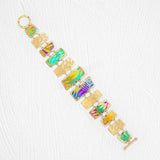 The Mahana Bracelet - 12 Rectangular Panels by Holly Yashi features rectangular, multicolored enamel panels and textured gold links, creating a vibrant tropical accessory beautifully displayed on a white textured background.