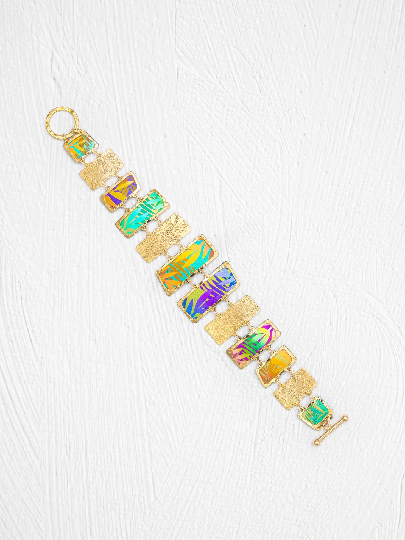 The Mahana Bracelet - 12 Rectangular Panels by Holly Yashi features rectangular, multicolored enamel panels and textured gold links, creating a vibrant tropical accessory beautifully displayed on a white textured background.